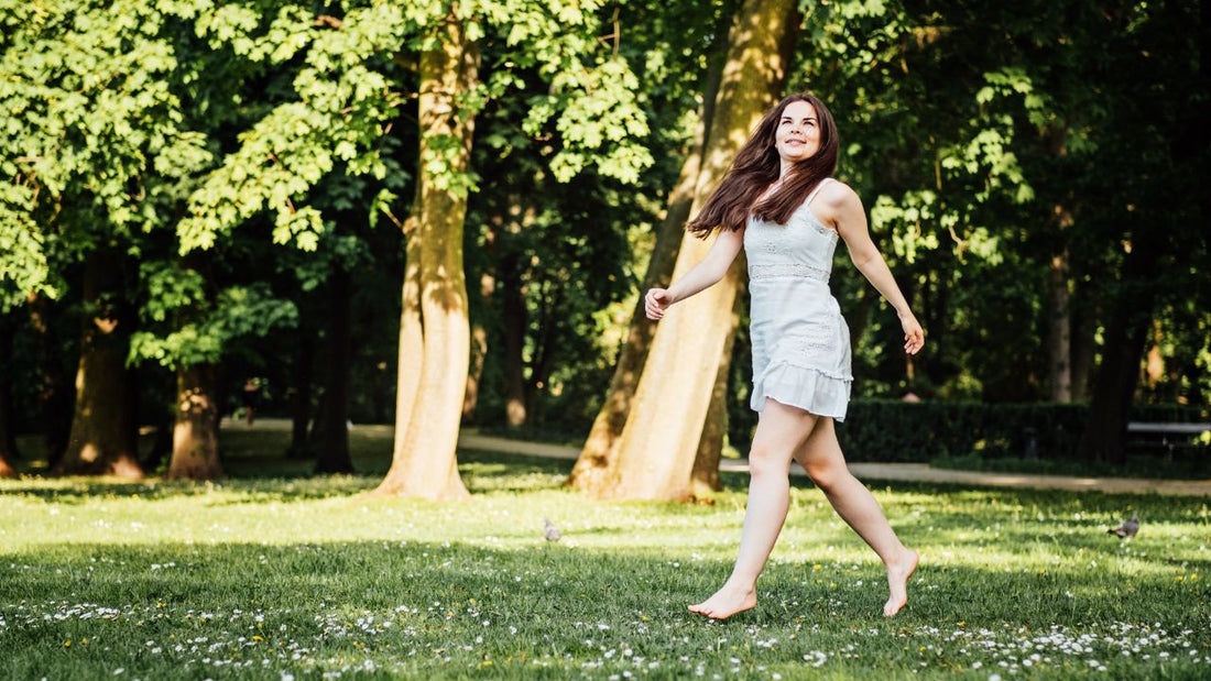 A woman joyfully walking barefoot on grass among trees, embodying the benefits of grounding.