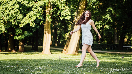 A woman joyfully walking barefoot on grass among trees, embodying the benefits of grounding.