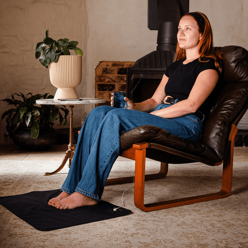 Woman sitting in a brown leather chair holding a blue mug in a cozy room with her feet on a grounding mat